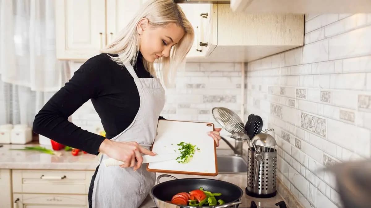 Žena v zástěře přidává nakrájenou cibulku do pánve se zeleninou