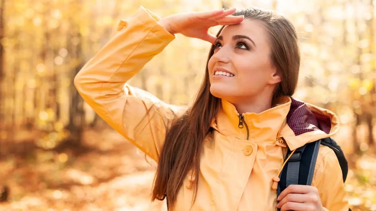 Žena ve žluté bundě s batohem se dívá do slunce v podzimním lese